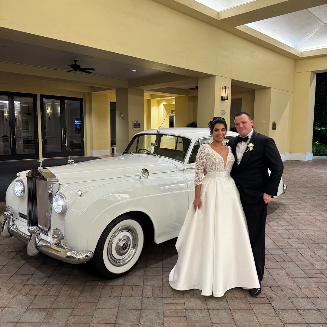 The Club at Grandezza Estero FL Wedding Transportation — Southerland Wedding Couple with Rolls Royce Silver Cloud II Bride in a lace gown and groom in a black tuxedo standing beside a vintage white Rolls Royce Silver Cloud II beneath the porte cochere at The Club at Grandezza in Estero, Florida