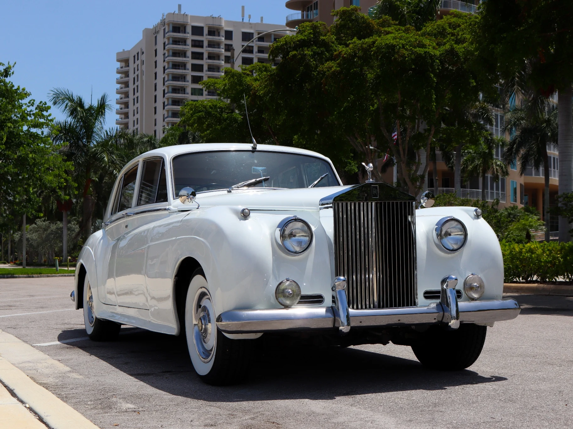 Front view of vintage white 1960 Rolls-Royce Silver Cloud II wedding transportation in Naples Florida with palm trees and coastal high-rise backdrop Front view of vintage white 1960 Rolls-Royce Silver Cloud II wedding transportation in Naples Florida with palm trees and coastal high-rise backdrop