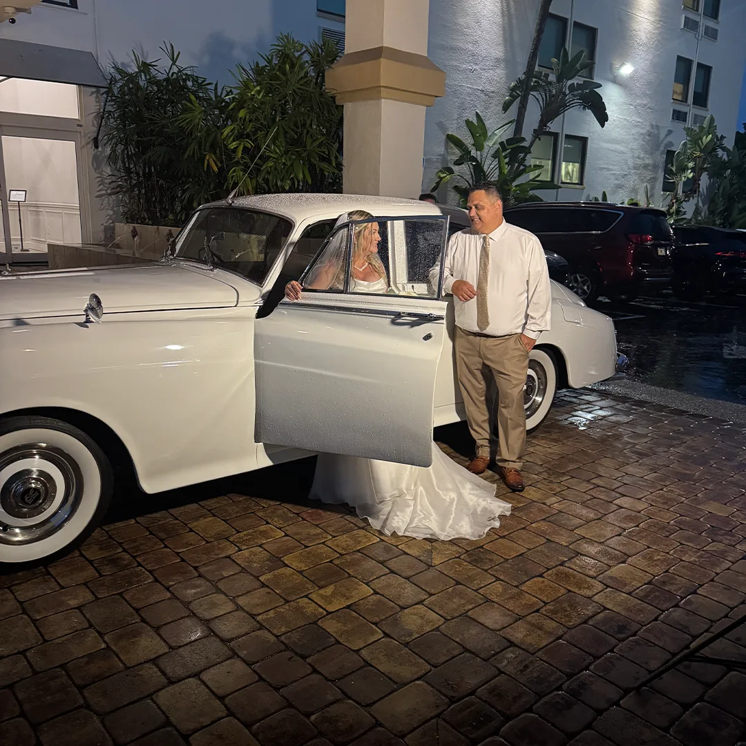Bayfront Inn Naples FL Wedding Transportation — Robbins Wedding Bride at the Rolls Royce Door Bride in a strapless gown stepping from the open door of a vintage white Rolls Royce Silver Cloud II on a wet brick driveway at the Bayfront Inn in Naples, Florida at night while a groomsman in a tan suit stands beside the car