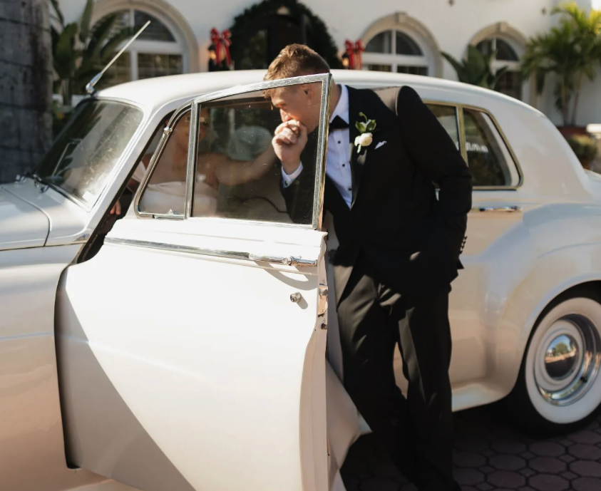 Groom kissing bride's hand beside vintage Rolls-Royce Silver Cloud wedding car in Naples Florida